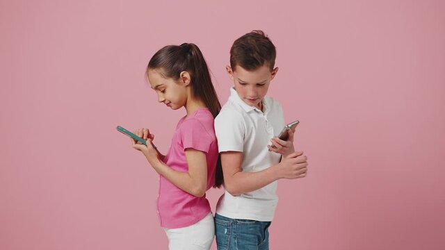 Kids and gadget addiction. Little sister and brother standing back to back and browsing on mobile phones