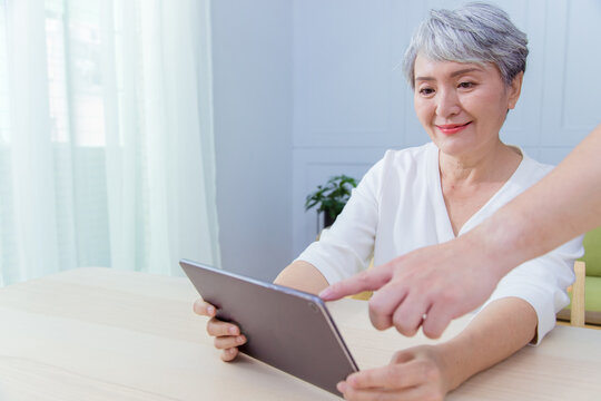 Young Friend Teaching Older Woman To Use Computer Tablet , Pointing At Screen, Elder Woman Asking Questions About Mobile Device. Elderly And Technology Concept.