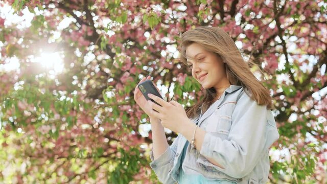 Hipster woman typing by mobile phone outdoors. Cheerful girl with smartphone in park on a background of sakura trees. Smiling lady holding cellphone in hands outside.