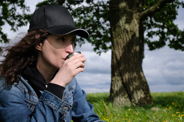 The girl is having lunch in the wild. Thermos, coffee, cheese and slate sandwich. Sunlight, trees, green grass, flowers, blue sky and rain clouds. Travel outside the city.