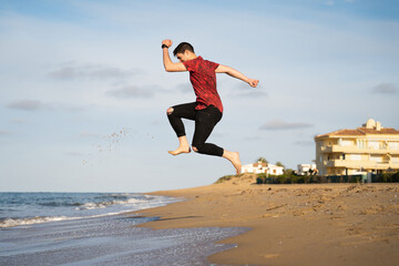 Man jumping on the seashore on a sunny day. Caucasian with long trousers and short-sleeved shirt. Shadow can be seen on the golden sand.