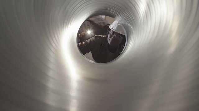 Chest-up view through pipe of young female African worker wearing white hard hat and transparent safety goggles, using flashlight on smartphone, looking inside