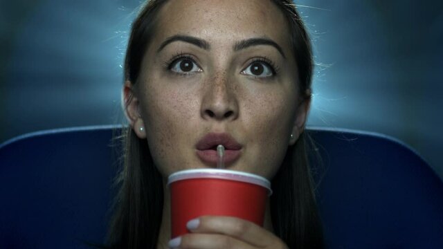 Captivated Young Woman At The Movie Theatre Sipping Her Drink With Projector Light Behind Her.