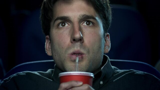 Closeup of a young man taking a sip of his drink while watching a movie at the movie theatre.