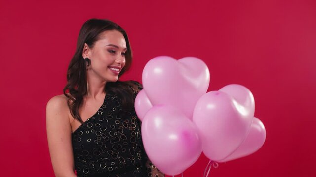 flattered woman in black dress receiving pink balloons isolated on red