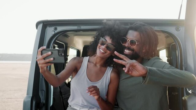 The smiling couple of Africans making selfie on the phone while sitting in the trunk of a car in the nature