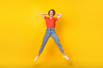 Fototapeta na wymiar Full size photo of cool brunette optimistic lady jump hands head wear red t-shirt jeans sneakers isolated on yellow color background