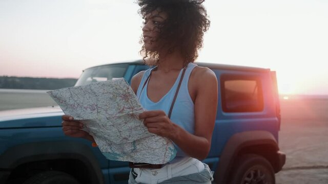 The African woman standing with a map in her hands near the car in front of the sunset on the sand