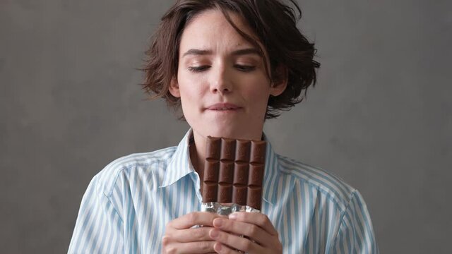 A close-up view of a woman with short hair holding a chocolate bar in her hands and biting off a piece with pleasure in a gray studio