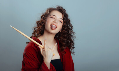 Rock and roll music concept. Cute caucasian young woman shows her tongue and and holds drumsticks in his hands on a gray-blue studio background. Free space for advertisement.