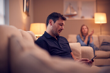 Evening Shot Of Couple On Sofa At Home With Man Looking At Mobile Phone And Unhappy Woman Behind