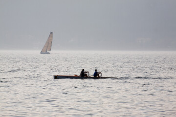 A sweep boat sails in the waters of a lake in northern Italy with two rowers and a sailboat is...
