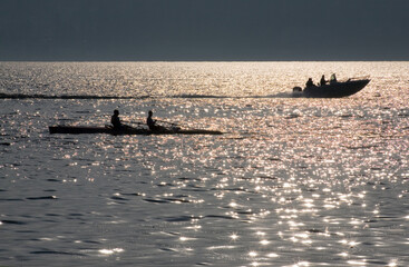A boat with two rowers and a motorboat sail on the calm waters of the sea illuminated by the setting sun in backlight