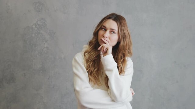 A pensive woman in hoodie looking to the sides and thinking about something while standing in a gray studio