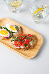 Toasted sandwiches white bread with cream cheese, vegetables: cherry tomatoes, cucumbers, eggs, parsley. Healthy vegetarian breakfast idea. Served with fresh lemon water, light background, copy space