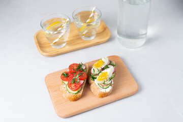 Toasted sandwiches white bread with cream cheese, vegetables: cherry tomatoes, cucumbers, eggs, parsley. Healthy vegetarian breakfast idea. Served with fresh lemon water, light background, copy space