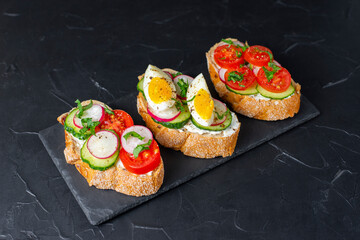 Delicious toasted sandwiches white bread with cream cheese, vegetables: cherry tomatoes, cucumbers, eggs, parsley. Healthy vegetarian breakfast idea. Served on black stone board, dark background