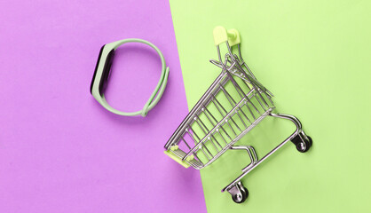 Mini shopping trolley with smart bracelet on pink green pastel background. Modern gadgets. Top view