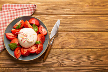 Italian salad with fresh tomatoes, burrata cheese, basil and olive oil served on ceramic plate on rustic wooden background from above, healthy snack or lunch with space for text