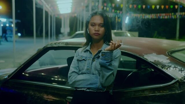 A girl smokes in front of a rusty classic car while typing a message late at night