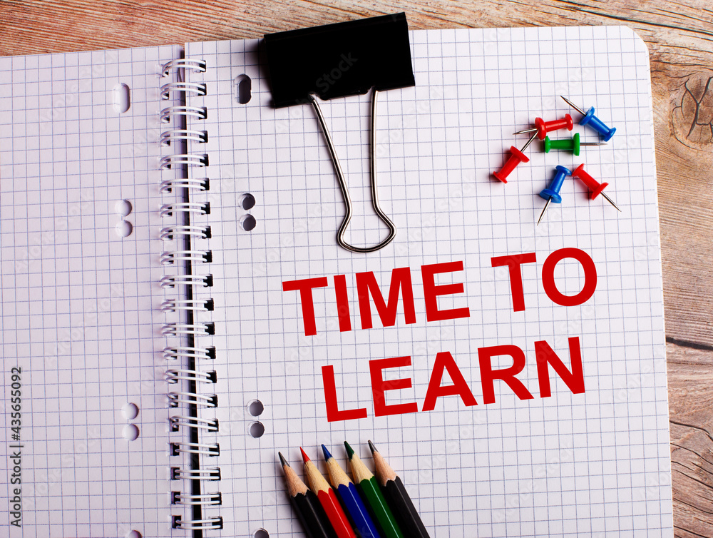 Poster the words time to learn is written in a notebook near multi-colored pencils and buttons on a wooden 