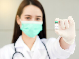 An Asian female doctor or nurse with a surgical mask is holding a vaccine bottle in her hand, focusing on the vaccine bottle in the foreground.