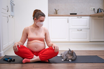Playing sports during pregnancy in the coronavirus pandemic. Pregnant woman does fitness workout in home kitchen in face medical mask