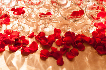 Fototapeta na wymiar The red rose petals are spilled in and around the wine goblet on the golden banquet table, accompanied by the light reflecting the dazzling light pattern, the perspective taken from top to bottom
