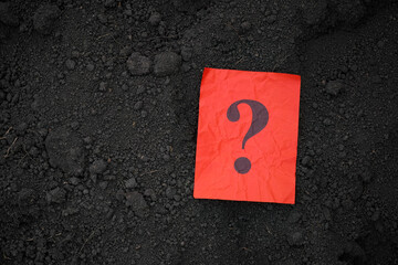 A red paper note with a question mark on it lying on soil