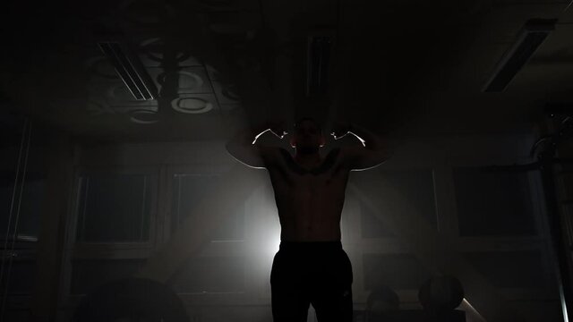 Bodybuilder flexing his muscles against the light in a dark gym.