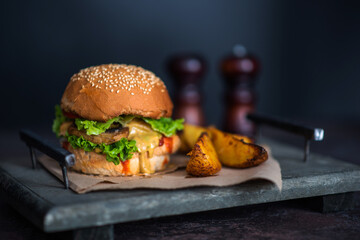 Tasty grilled homemade beef burger, tomato, cheese, cucumber and lettuce with french fries on a wooden background. Burger on the board on a dark background.