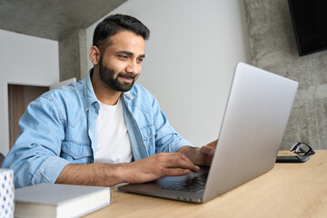 Young happy Indian businessman using computer working remotely from modern home office. Latin teacher having virtual training on laptop typing watching distant online webinar during quarantine.