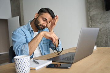 Young indian eastern tired exhausted business man rubbing eyes sitting in modern home office with laptop on desk. Overworked burnout academic Hispanic student with glasses in hand feeling eyestrain. - obrazy, fototapety, plakaty