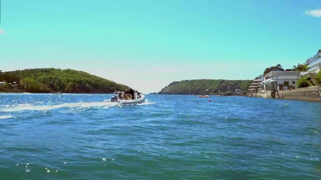 Speed boat zooms past camera from left side of shot to right at Salcombe harbour out through the estuary towards the sea. Bright blue sky in the background on a summer day in South West Devon