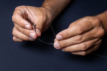 Fishing line and hook being handled by the hands of a brown man demonstrating how to thread the line correctly on a black and dark background. Used for fishing and family leisure.