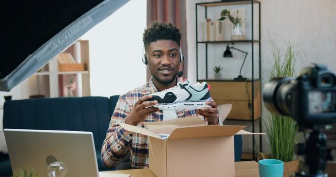 Blogging,shopping and presentation concept where likable satisfied professional black-skinned male blogger recording unpacking process of new-buyed sneakers on camera