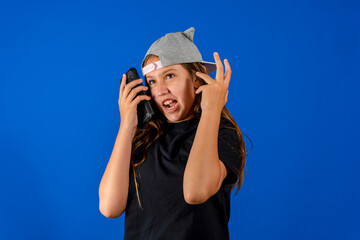 Obraz na płótnie Canvas Pretty preteen girl in a cap talking pissed off on the cellphone isolated on blue studio background