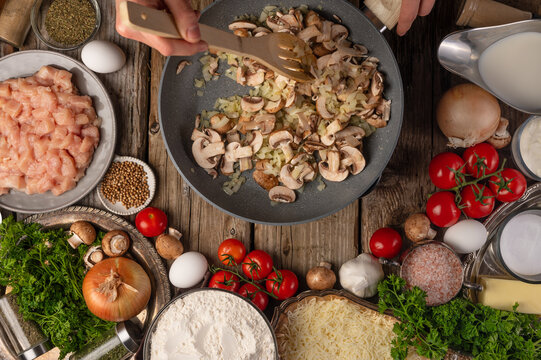 Many ingredients are laid out on a wooden table. Mushrooms are cooked in a pan, cut into pieces. The cook is stirring the mushrooms with a wooden spoon. View from above. Color image. High angle view.