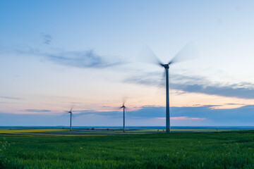 Dramatic landscape of wind power stations in a wind farm with propellers moving against the background of blue and orange sky during sunset.