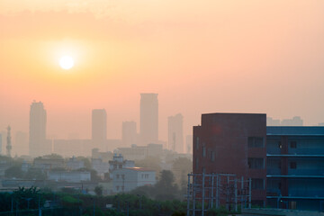 wide shot of gurgaon delhi cityscape showing a brick building and houses in the foreground and tall buildings with many floors in the fog haze obsured distance against the sun rising