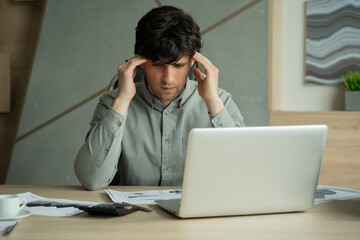 Frustrated man while sitting at his working place in office. Exhausted young man with laptop in office