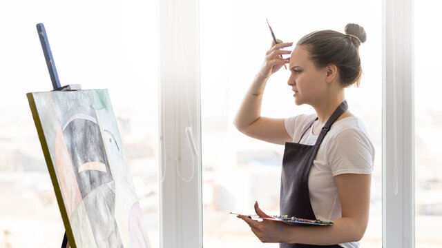 Thoughtful female artist. Creative process. Find inspiration. Art studio. Puzzled woman touching forehead holding brush and palette paints looking on easel with artwork light interior.