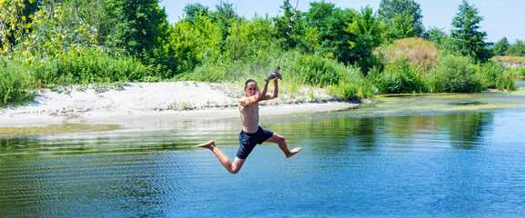 Boy teenager swings on a rope in a funny pose and ready to jump into the water on sunny summer day. Beautiful landscape of a river with green banks. Concept of healthy lifestyle