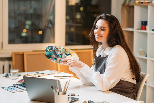 Online education. Female artist. Color array. Art school. Painting blog. Happy inspired woman looking laptop showing palette with paints splashes in light studio interior.
