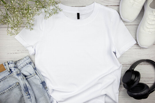 White womens cotton Tshirt mockup with flowers, jeans, sneakers and black headphones on wooden background. Design t shirt template, print presentation mock up. Top view flat lay. 