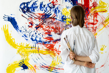 Modern art. Painting inspiration. Imagination talent. Back view of female painter with paintbrushes enjoying looking at colorful smeared handprint abstract design artwork on white wall.