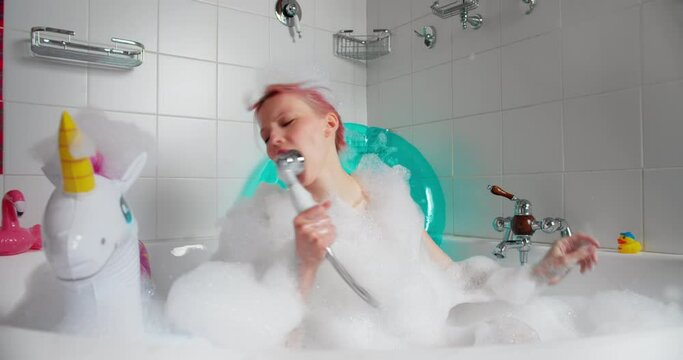 Funny woman sits in bath filled with foam, holds shower singing songs. Female in foam from head to toe in the bath fooling around
