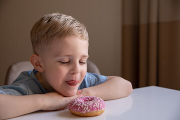 The concept is useless food. A little boy with blond hair eats a pink doughnut at home. He's happy.