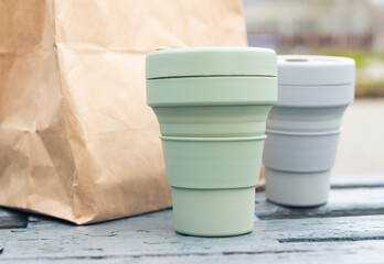 Two collapsible eco cups and craft parcel on city park bench. Reusable sustainable silicon mugs for takeaway tea or coffee.