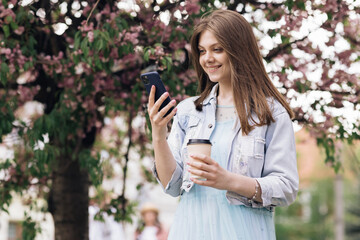 Woman with charming Smile walking in the Park and Using Mobile Phone Wearing Stylish Outfit. Girl Using Smartphone and Drinking Coffee. Communication, online shopping, social network concept